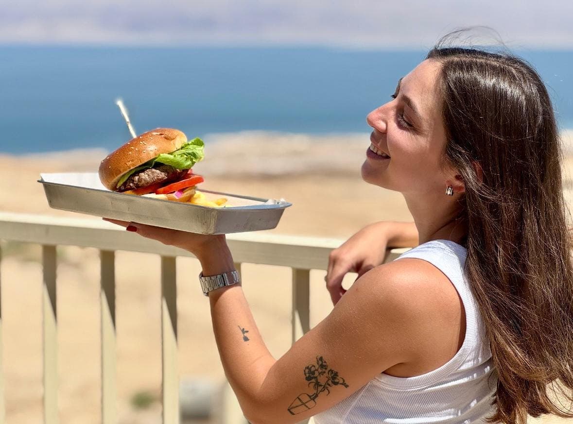 אורחת מחזיקה מגש עם המבורגר וצ'יפס על רקע ים המלח